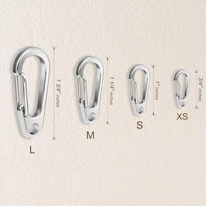May include: Four silver-colored metal carabiner clips arranged by size from left to right, labeled L, M, S, and XS. The sizes are indicated with vertical measurement lines showing 1 3/4 inches, 1 1/4 inches, 1 inch, and 3/4 inches respectively.