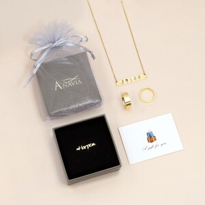 May include: A gray gift box with the word "Anavia" printed on it, a small gray box with a gold ring that says "Harper" inside, a gold necklace with a bar pendant that says "1999", a gold ring, and a white card with a gift illustration and the text "A gift for you".