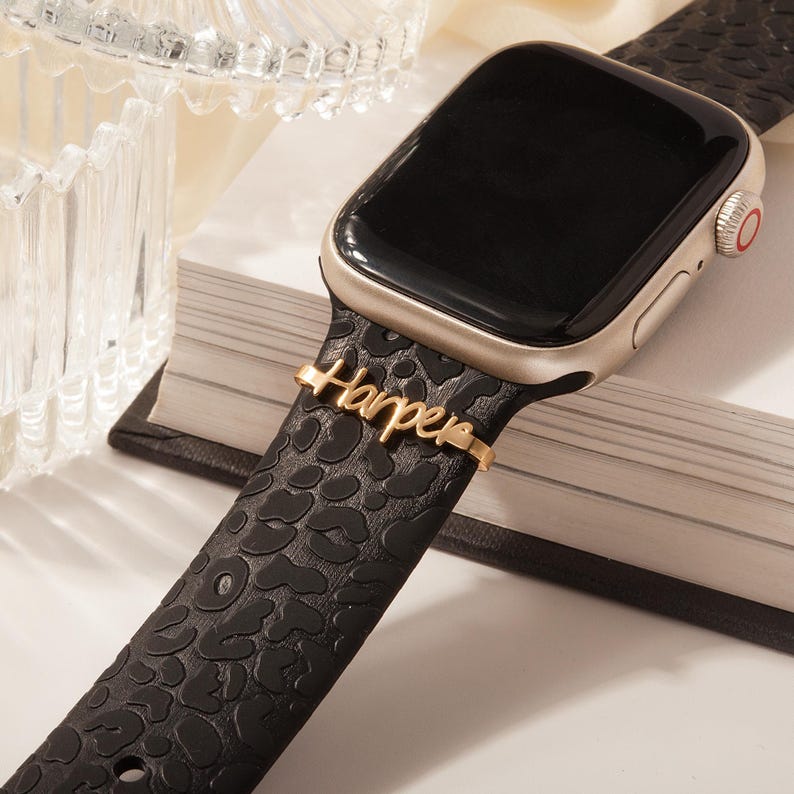 May include: Black and gold Apple Watch with a leopard print band. The band has a gold charm with the name "Harper" engraved on it.