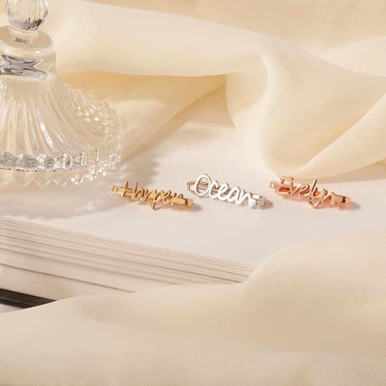 May include: Three gold rings with the names "Harper", "Ocean", and "Evelyn" engraved on them. The rings are on a white surface.