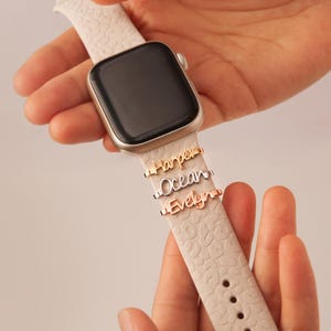 May include: A white Apple Watch with a white band featuring three personalized name charms. The charms are in silver, gold, and rose gold and spell out "Harper", "Ocean", and "Evelyn".