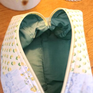 May include: Open, zippered cosmetic bag with a yellow zipper and a green interior. The exterior features a patchwork design with lemon and floral patterns in yellow, blue, and white. The bag is open, revealing the green lining.