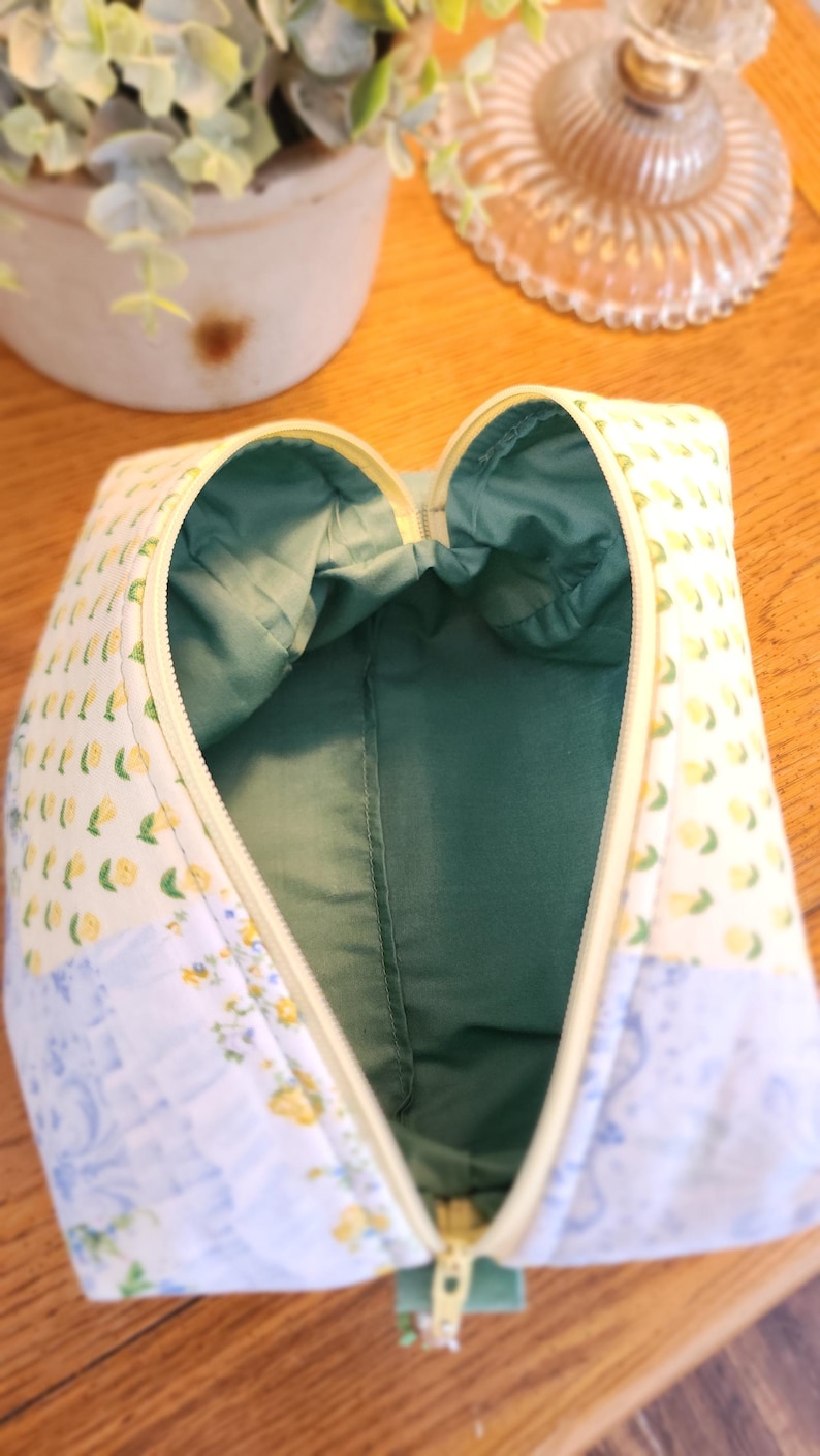May include: Open, zippered cosmetic bag with a yellow zipper and a green interior. The exterior features a patchwork design with lemon and floral patterns in yellow, blue, and white. The bag is open, revealing the green lining.