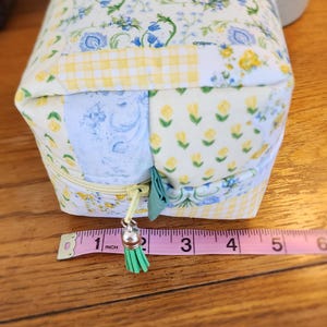 May include: A rectangular fabric pouch with a zipper closure. The pouch is approximately 5 inches long. It features a patchwork design with floral and gingham patterns in blue, yellow, and white. A green tassel hangs from the zipper.