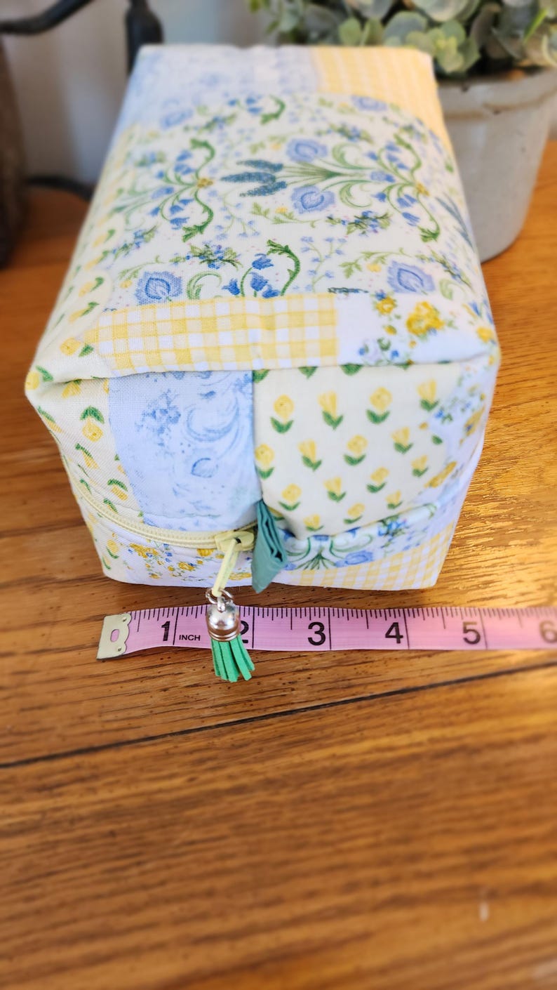 May include: A rectangular fabric pouch with a zipper closure. The pouch is approximately 5 inches long. It features a patchwork design with floral and gingham patterns in blue, yellow, and white. A green tassel hangs from the zipper.