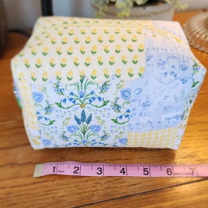 May include: A quilted fabric cosmetic bag with a floral pattern. The bag features a patchwork design with yellow, blue, and white fabrics. The bag is approximately 6 inches wide.