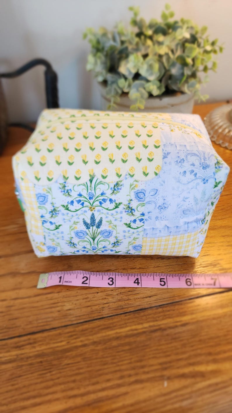 May include: A quilted fabric cosmetic bag with a floral pattern. The bag features a patchwork design with yellow, blue, and white fabrics. The bag is approximately 6 inches wide.