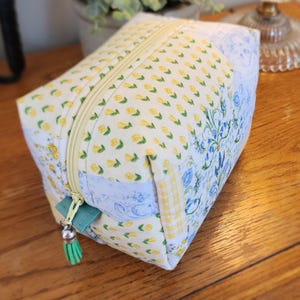 May include: A rectangular cosmetic bag with a yellow floral pattern, a light green zipper pull, and a matching tassel. The bag features a mix of floral and patterned fabrics, with a light blue floral design on one side.