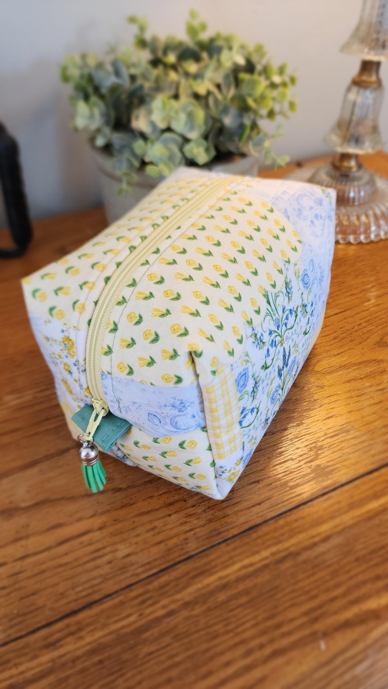 May include: A rectangular cosmetic bag with a yellow floral pattern, a light green zipper pull, and a matching tassel. The bag features a mix of floral and patterned fabrics, with a light blue floral design on one side.