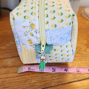 May include: A small, rectangular fabric pouch with a yellow zipper and a green tassel. The pouch is made of patchwork fabric with floral and gingham patterns. The pouch is approximately 5 inches long.