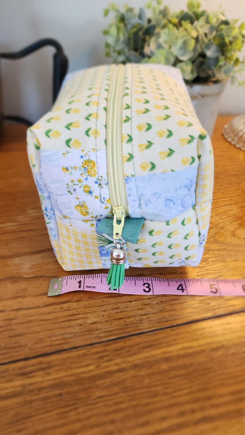 May include: A small, rectangular fabric pouch with a yellow zipper and a green tassel. The pouch is made of patchwork fabric with floral and gingham patterns. The pouch is approximately 5 inches long.