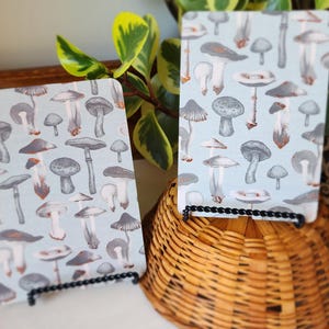 May include: Two rectangular cards featuring a repeating pattern of various mushrooms in shades of gray and brown against a light blue background. Each card is displayed on a small black wire stand, with one card positioned slightly lower than the other.