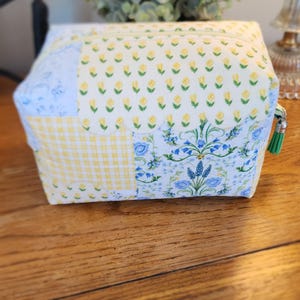 May include: A patchwork cosmetic bag with a zipper closure and a green tassel. The bag features a variety of floral and gingham patterns in yellow and blue. The bag is sitting on a wooden surface.