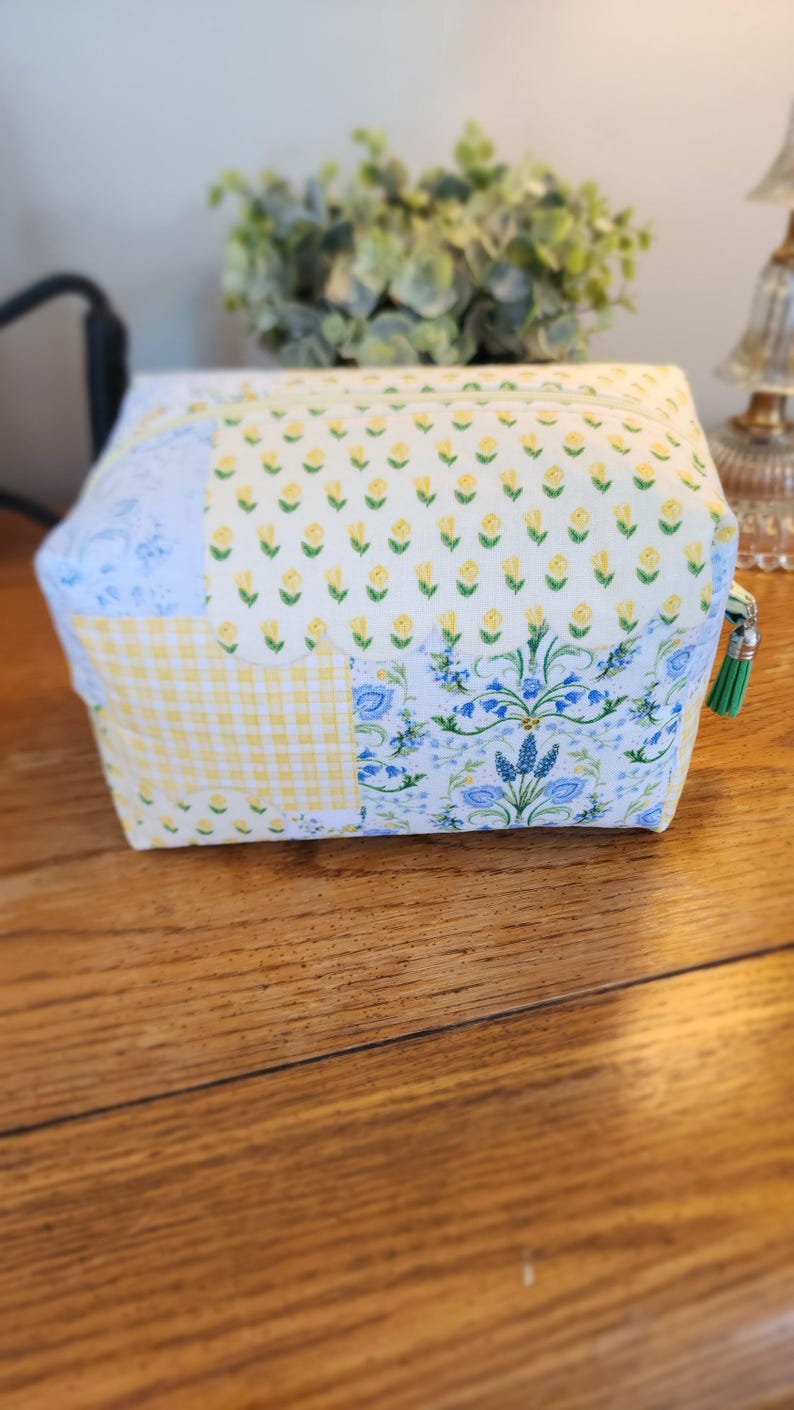 May include: A patchwork cosmetic bag with a zipper closure and a green tassel. The bag features a variety of floral and gingham patterns in yellow and blue. The bag is sitting on a wooden surface.