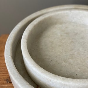 May include: Two white marble bowls, one nested inside the other. The bowls are round and have a smooth, polished surface.