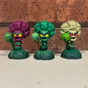 May include: Three quirky figurines, each a vegetable-inspired character. One is purple, one green, and one off-white, all with leafy bases. They hold green orbs and have yellow or red eyes. A fun, collectible item.