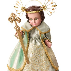 Könnte beinhalten: Detaillierte Puppe einer religiösen Figur, die ein hellgrünes und goldenes Kleid mit aufwendiger Stickerei trägt. Die Puppe hat einen goldenen Heiligenschein und hält ein goldenes Kreuz. Das Gesicht der Puppe hat grüne Augen und braunes Haar.
