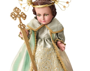 May include: A detailed doll of a religious figure, wearing a light green and gold dress with intricate embroidery. The doll has a gold halo and holds a gold cross. The doll's face has green eyes and brown hair.