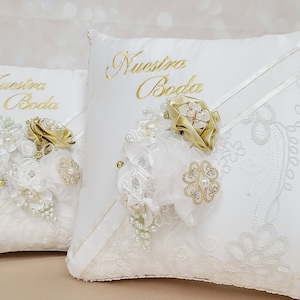 May include: Two white satin pillow covers with lace trim and gold embroidery. The larger pillow has the words "Nuestra Boda" embroidered on it. Both pillows have a white and gold floral embellishment.