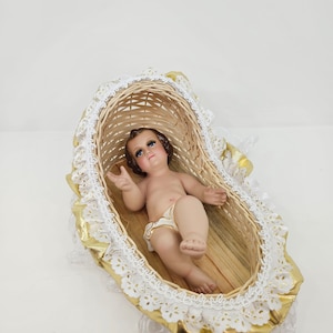 K&ouml;nnte beinhalten: Eine Babyfigur mit braunen Haaren und blauen Augen liegt in einem kleinen, ovalen geflochtenen Weidenkorb. Der Korb ist mit einer wei&szlig;en Spitzenborte und einem goldfarbenen Stoffrand verziert, was ihm das Aussehen einer Wiege verleiht.