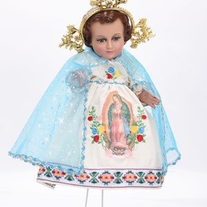 Puede incluir: Una mu&ntilde;eca de porcelana blanca vestida con un vestido blanco con ribete bordado azul y blanco. El vestido presenta una imagen impresa de la Virgen Mar&iacute;a. La mu&ntilde;eca lleva una capa de tul azul con purpurina plateada y una corona dorada con pedrer&iacute;a.