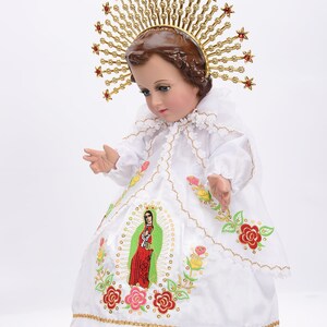 Puede incluir: Una mu&ntilde;eca de porcelana blanca vestida con un vestido blanco con ribetes dorados y flores bordadas. La mu&ntilde;eca lleva una corona dorada con joyas rojas y una corona de espinas en la cabeza. La mu&ntilde;eca mira hacia abajo y tiene una expresi&oacute;n serena.