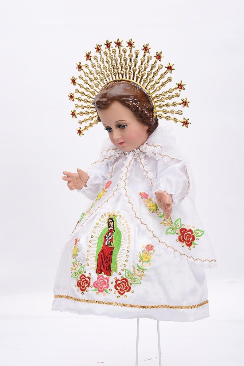 Puede incluir: Una mu&ntilde;eca de porcelana blanca vestida con un vestido blanco con ribetes dorados y flores bordadas. La mu&ntilde;eca lleva una corona dorada con joyas rojas y una corona de espinas en la cabeza. La mu&ntilde;eca mira hacia abajo y tiene una expresi&oacute;n serena.