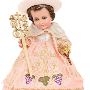 May include: A porcelain doll dressed in a pink and gold gown with embroidered grapevines and a cross. The doll is wearing a white hat and holding a wooden cross with grapevines.
