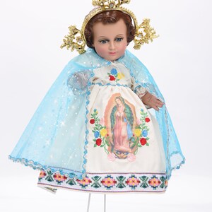 Puede incluir: Una mu&ntilde;eca de porcelana blanca vestida con un vestido blanco con una capa azul y una corona dorada. El vestido tiene una imagen bordada de la Virgen Mar&iacute;a y detalles florales. La mu&ntilde;eca est&aacute; en un soporte blanco.