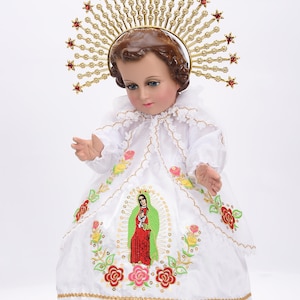 Puede incluir: Una mu&ntilde;eca de porcelana blanca vestida con un vestido blanco bordado con flores y una representaci&oacute;n de la Virgen Mar&iacute;a. La mu&ntilde;eca lleva una corona dorada con joyas rojas y tiene un halo.