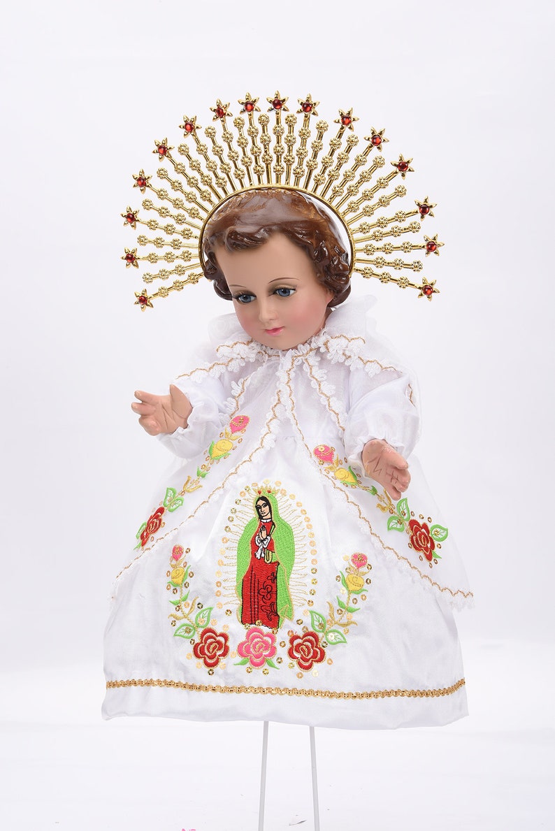 Puede incluir: Una mu&ntilde;eca de porcelana blanca vestida con un vestido blanco bordado con flores y una representaci&oacute;n de la Virgen Mar&iacute;a. La mu&ntilde;eca lleva una corona dorada con joyas rojas y tiene un halo.