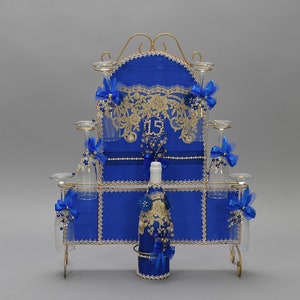May include: A royal blue and gold decorative display with a champagne bottle, glasses, and candle holders. The display features a gold metal frame, blue fabric, and ornate gold lace. The number "15" is displayed on the front.