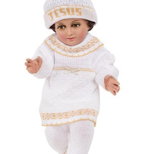 May include: A white knitted baby doll outfit with gold trim and a matching white knitted hat with the word "JESUS" embroidered in gold.