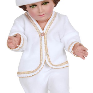 Könnte beinhalten: Eine weiße Puppe in einem passenden Outfit, einschließlich Hut, Jacke und Hose. Die Kleidung ist mit goldfarbenen Details verziert. Die Puppe hat grüne Augen und braunes Haar.