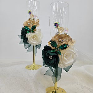 Könnte beinhalten: Zwei Champagnerflöten mit goldenen Rändern, verziert mit weißen und smaragdgrünen Stoffblumen, goldenem Band und funkelnden Strasssteinen. Die Flöten stehen auf einer weißen, glitzernden Oberfläche.
