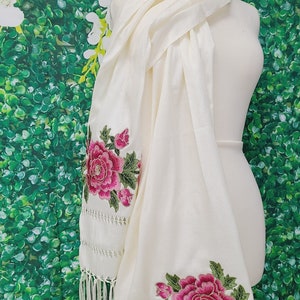 May include: Cream-colored scarf with embroidered pink floral design and fringe. The scarf is draped over a mannequin. The floral embroidery features large, detailed flowers with green leaves.