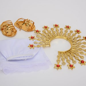 Puede incluir: Una corona dorada con joyas rojas, una prenda de tela blanca y dos sandalias doradas. La corona es de estilo halo con una banda circular y puntos radiantes.