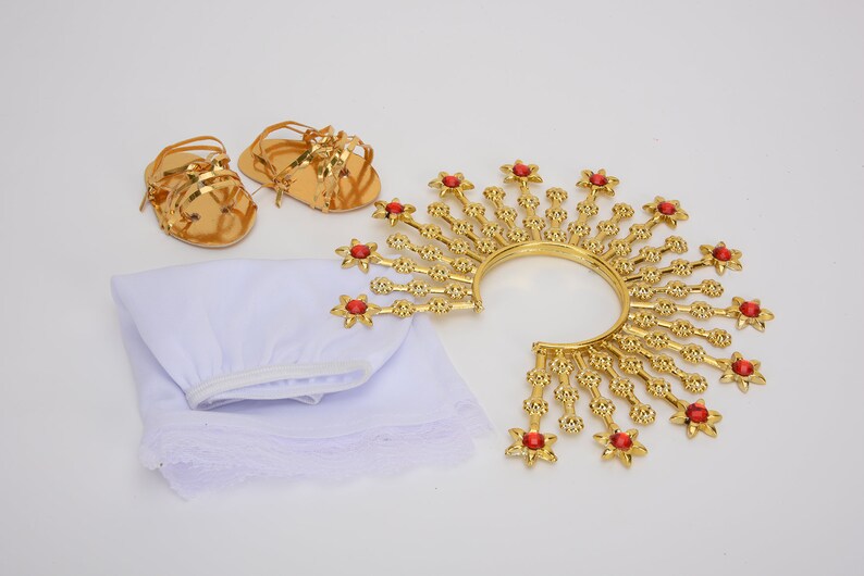 Puede incluir: Una corona dorada con joyas rojas, una prenda de tela blanca y dos sandalias doradas. La corona es de estilo halo con una banda circular y puntos radiantes.