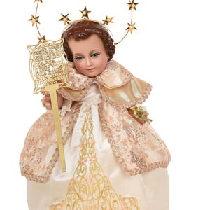 May include: A porcelain doll of the Infant Jesus of Prague, dressed in a white and gold gown with a lace trim. The doll is holding a golden scepter with a prayer card attached. The doll is wearing a gold halo with stars.
