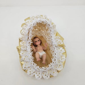 K&ouml;nnte beinhalten: Eine kleine Babyfigur, die in einer geflochtenen Korbwiege liegt. Die Wiege ist mit einer wei&szlig;en Lochspitzenborte und gl&auml;nzenden goldfarbenen Stoffr&uuml;schen verziert. Das Baby hat braune Haare, bemalte Gesichtsz&uuml;ge und tr&auml;gt eine wei&szlig;e Windel.