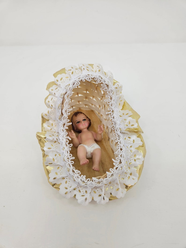 K&ouml;nnte beinhalten: Eine kleine Babyfigur, die in einer geflochtenen Korbwiege liegt. Die Wiege ist mit einer wei&szlig;en Lochspitzenborte und gl&auml;nzenden goldfarbenen Stoffr&uuml;schen verziert. Das Baby hat braune Haare, bemalte Gesichtsz&uuml;ge und tr&auml;gt eine wei&szlig;e Windel.