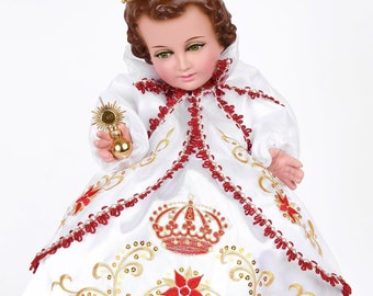 Puede incluir: Un vestido blanco bordado con detalles rojos y dorados. El vestido presenta una corona y diseños florales. Una pequeña muñeca de niño lleva el vestido y una corona dorada.