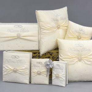 Könnte beinhalten: Ein Set aus cremefarbenen Satin-Hochzeits-Schmuckkästen und Kissen mit silbernen Stickereien und Verzierungen. Die Kästen und Kissen zeigen den Text "Nuestra Boda" und ein Paar Ringe.