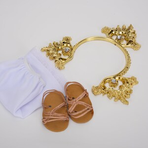 Puede incluir: Un conjunto de ropa de beb&eacute; blanca con una diadema dorada y sandalias marrones. La diadema est&aacute; decorada con pedrer&iacute;a.