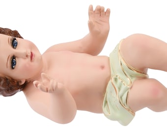 May include: A ceramic figurine of a baby Jesus lying on its back, wearing a white and green swaddling cloth. The baby has brown hair, blue eyes, and a peaceful expression.