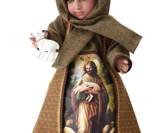 Puede incluir: Muñeca religiosa que representa una figura con una túnica con capucha marrón, sosteniendo un cordero blanco. La túnica de la muñeca presenta una imagen impresa de Jesús sosteniendo un cordero. La muñeca mide unos 30 cm de altura.