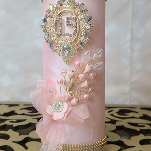 May include: A pink candle with a gold and rhinestone embellishment featuring the letter "L" and a pink flower with rhinestones. The candle is decorated with gold and rhinestone trim.