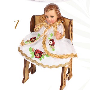 Puede incluir: Una peque&ntilde;a mu&ntilde;eca de porcelana vestida con un vestido blanco con ribetes dorados y bordados de flores rojas. La mu&ntilde;eca est&aacute; sentada en una silla de madera.