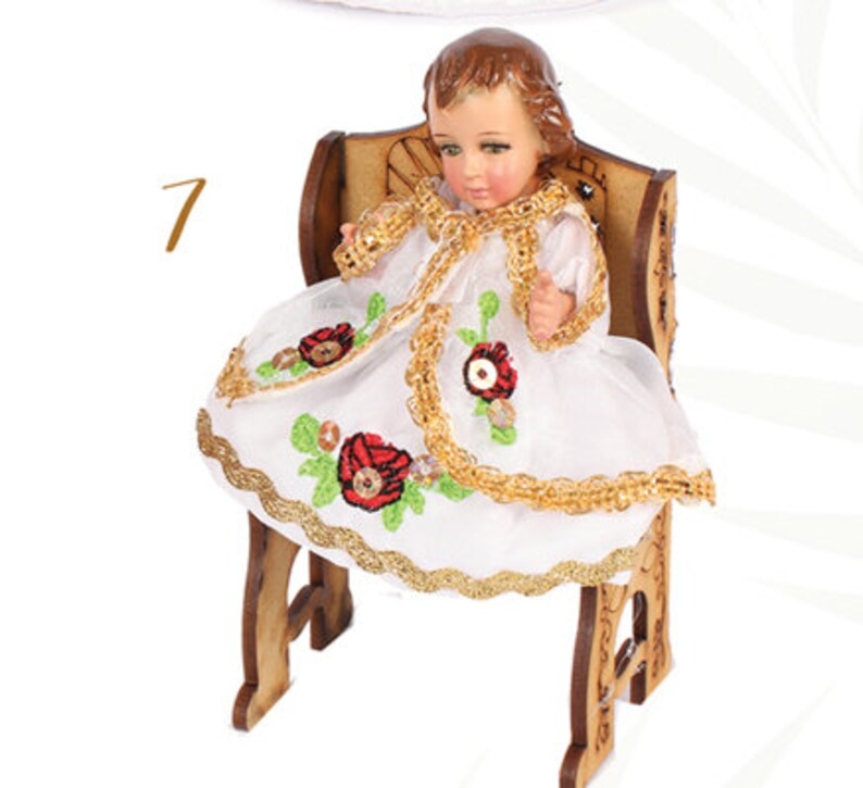Puede incluir: Una peque&ntilde;a mu&ntilde;eca de porcelana vestida con un vestido blanco con ribetes dorados y bordados de flores rojas. La mu&ntilde;eca est&aacute; sentada en una silla de madera.