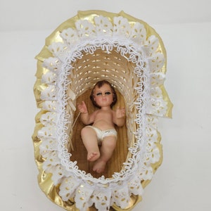 K&ouml;nnte beinhalten: Eine kleine Babyfigur mit braunen Haaren und gr&uuml;nen Augen, bekleidet mit einer wei&szlig;en Windel, liegt in einer geflochtenen Korbwiege. Die Wiege ist mit einer ger&uuml;schten wei&szlig;en Spitzenborte und einem gl&auml;nzenden goldenen Stoffrand verziert.